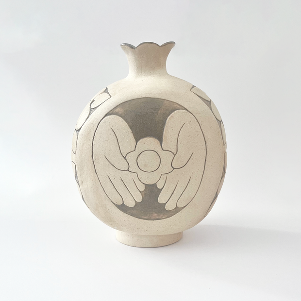 Cream colored ceramic vase with a very round body and a small fluted top, almost resembling a pomegranate in shape. On the back is a design of a person's hands holding a flower.