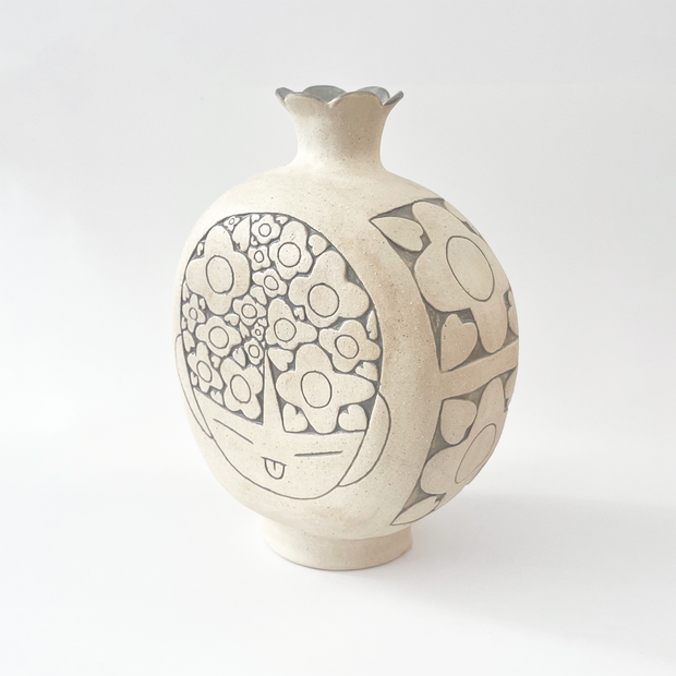  Cream colored ceramic vase with a very round body and a small fluted top, almost resembling a pomegranate in shape. On the front is a design of a person's head with floral patterned hair.