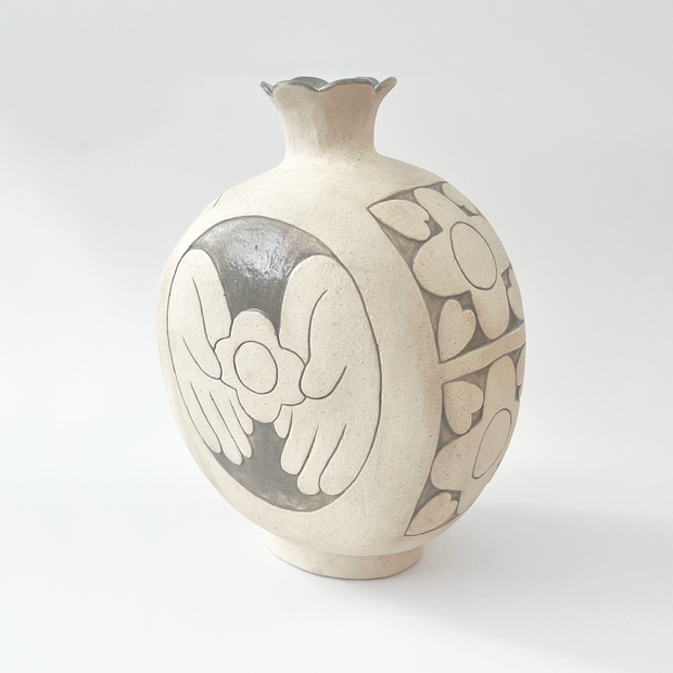 Cream colored ceramic vase with a very round body and a small fluted top, almost resembling a pomegranate in shape. On the back is a design of a person's hands holding a flower.