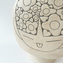 Cream colored ceramic vase with a very round body and a small fluted top, almost resembling a pomegranate in shape. On the front is a design of a person's head with floral patterned hair.