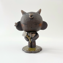 Dark brown ceramic sculpture of a character with a large head, large ears and a tiny body. They have horns and earring cuffs. They stand and hold a white flower with a thorny stem.
