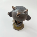 Dark brown ceramic sculpture of a character with a large head, large ears and a tiny body. They have horns and earring cuffs. They stand and hold a white flower with a thorny stem.