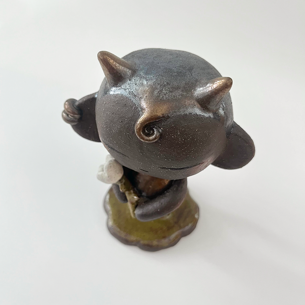 Dark brown ceramic sculpture of a character with a large head, large ears and a tiny body. They have horns and earring cuffs. They stand and hold a white flower with a thorny stem.
