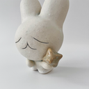 Small white ceramic sculpture of a bunny character with a calm, closed eye expression. It has a large head and a tiny body, holding a star.