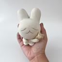 Hand holding a white ceramic sculpture of a bunny character with a calm, closed eye expression. It has a large head and a tiny body, holding a star.
