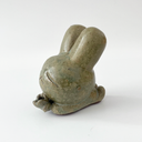 Small sage green ceramic sculpture of a bunny character with a giant head and a tiny body. It holds out a small tulip like flower in its hands.