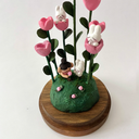 Clay sculpture enclosed in a glass cloche with a wood base of a grassy mound, with pink flowers growing with tall green, leafy stems. Small white rabbits sit in and around them and at in the grass a small girl smells a pink flower.