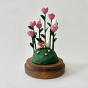 Clay sculpture enclosed in a glass cloche with a wood base of a grassy mound, with pink flowers growing with tall green, leafy stems. Small white rabbits sit in and around them and at in the grass a small girl smells a pink flower.