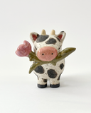 Ceramic figurine with a spotted clay finish of a cow with a flower in its mouth.