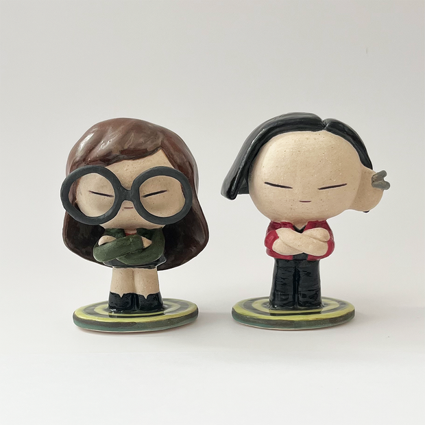 Ceramic sculpture of a stylized Daria from the show Daria - a girl with long brown hair, large glasses, a green coat and a black skirt with boots. She stands on a bullseye colored green, next to her companion Jane Lane.