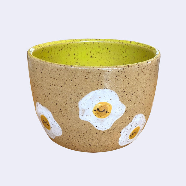 Ceramic bowl with spotted finishing and an earthy brown exterior and lime green interior. On the outside are painted on cartoon style eggs, with simple expressions.