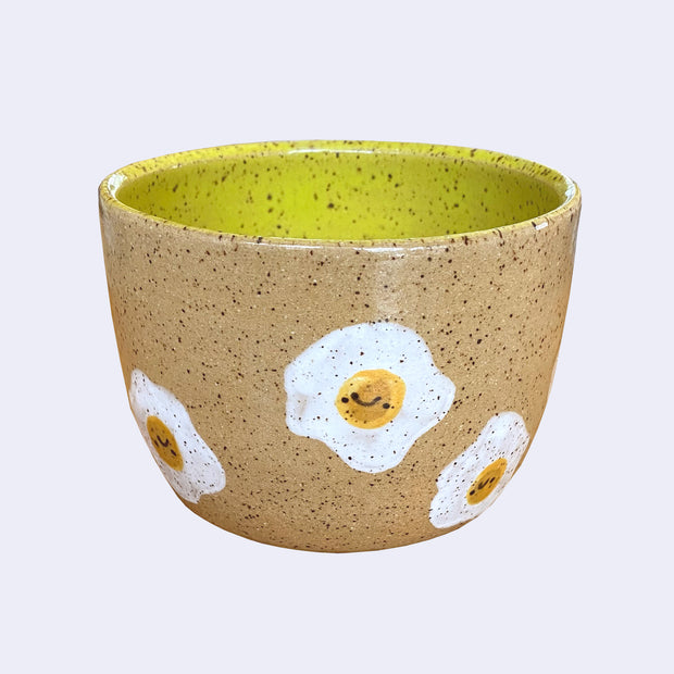 Ceramic bowl with spotted finishing and an earthy brown exterior and lime green interior. On the outside are painted on cartoon style eggs, with simple expressions.