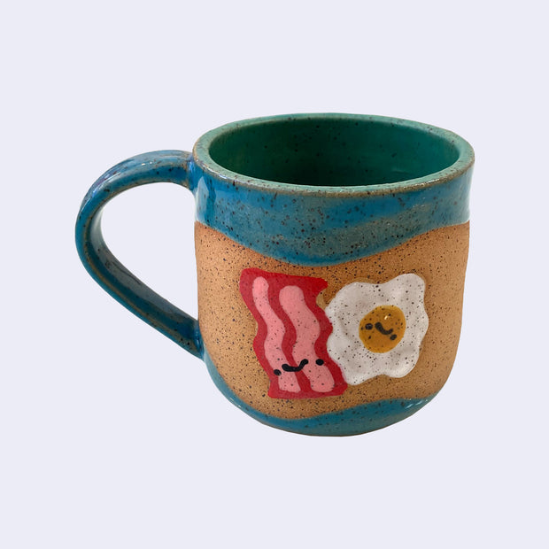 Ceramic mug with spotted finishing and an earthy brown exterior and bluish green interior. On the outside are painted on cartoon style bacon and eggs, with simple expressions.