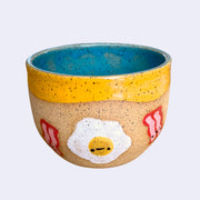Ceramic bowl with spotted finishing and an earthy brown exterior and teal blue interior. On the outside are painted on cartoon style eggs and bacon, with simple expressions.
