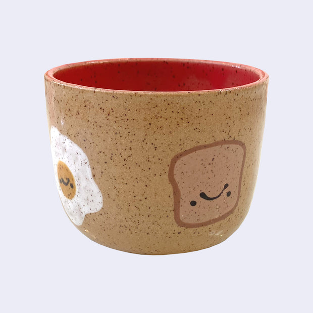 Ceramic bowl with spotted finishing and an earthy brown exterior and pinkish red interior. On the outside are painted on cartoon style eggs and toast, with simple expressions.
