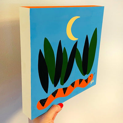 Solid color painting on panel of a blue background. A simply painted orange and black snake is at the bottom between blades of tall grass. A simplistic crescent moon hangs overhead.