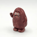 A small hand-painted ceramic figurine of a round, reddish-brown character with a white face, black eyes, and a red mouth, standing on a white background.