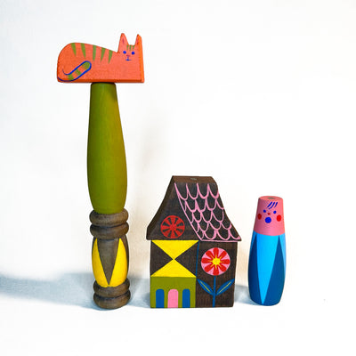 4 different small wooden sculptures, making a composition of a small house, a person, a post resembling a tree and an orange cat atop it.