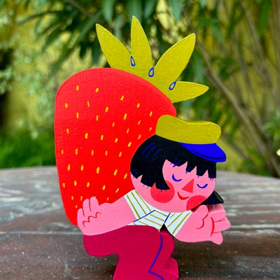 Wooden die cut sculpture of a woman with a short black bob cut wearing a green hat, striped shirt and burgundy pants. She hunches over and carries a very large strawberry on her back.