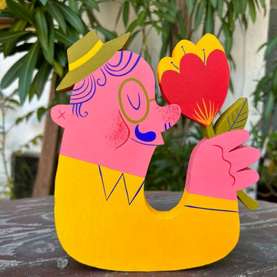 Die cut wooden sculpture of a pink man, facing off to the side, and holding a large flower raised up to his nose. Only his head and arm is visible. He wears a hat, glasses, and a closed eye smiling expression.