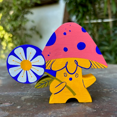 Die cut wooden sculpture of a yellow mushroom with a pink cap, with a small dazed expression, It holds a large circular white daisy with a blue outline, over its shoulder. 