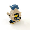 A hand-painted ceramic figurine of an onigiri character riding a skateboard, wearing a blue cap with yellow color accents, set against a plain white background.