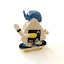 A hand-painted ceramic figurine of an onigiri character riding a skateboard, wearing a blue cap with yellow color accents, set against a plain white background.