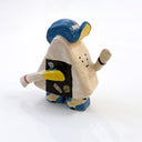 A miniature ceramic onigiri character, walking with its arms in full stride. It wears a blue and yellow cap, has yellow sleeves, yellow and blue sneakers and stickers on the side of its body.