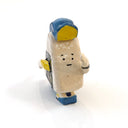A miniature ceramic onigiri character, walking with its arms in full stride. It wears a blue and yellow cap, has yellow sleeves, yellow and blue sneakers and stickers on the side of its body.