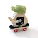 A miniature ceramic onigiri character, riding a skateboard, with its arms in full stride. It wears a green cap, has blue sleeves, blue and green sneakers and stickers on the side of its body.