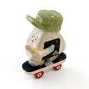 A miniature ceramic onigiri character, riding a skateboard, with its arms in full stride. It wears a green cap, has blue sleeves, blue and green sneakers and stickers on the side of its body.