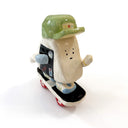 A miniature ceramic onigiri character, riding a skateboard, with its arms in full stride. It wears a green cap, has blue sleeves, blue and green sneakers and stickers on the side of its body.