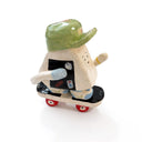 A miniature ceramic onigiri character, riding a skateboard, with its arms in full stride. It wears a green cap, has blue sleeves, blue and green sneakers and stickers on the side of its body.