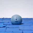 Carved wooden sculpture of a small blue colored moon, with a flat bottom. It has several small craters and a closed eye, calm facial expression. 
