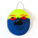 Sculpture of a flat clay mask with 3D eyes, nose and lips. It is half fluorescent yellow and half dark blue. It has red eyes and eyebrows and hangs from a chain.