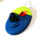 Sculpture of a flat clay mask with 3D eyes, nose and lips. It is half fluorescent yellow and half dark blue. It has red eyes and eyebrows and hangs from a chain.