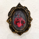 An ornate oval wall art piece with a distressed gold and black frame, featuring a red and black illustration of a kneeling winged figure on a textured white wall.