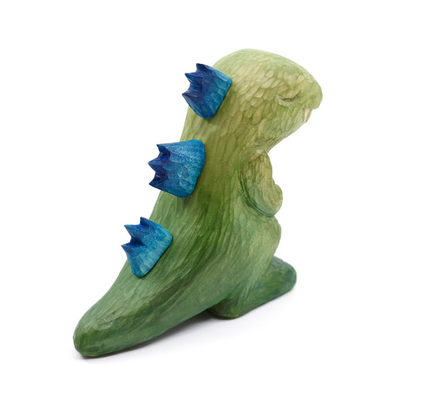 Whittled wooden sculpture of a green Godzilla with blue back spikes, modeled in a non aggressive, cute fashion. Its eyes are peacefully closed and its hands rest on its chest.