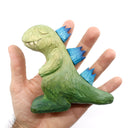 Whittled wooden sculpture of a green Godzilla with blue back spikes, modeled in a non aggressive, cute fashion. Its eyes are peacefully closed and its hands rest on its chest.