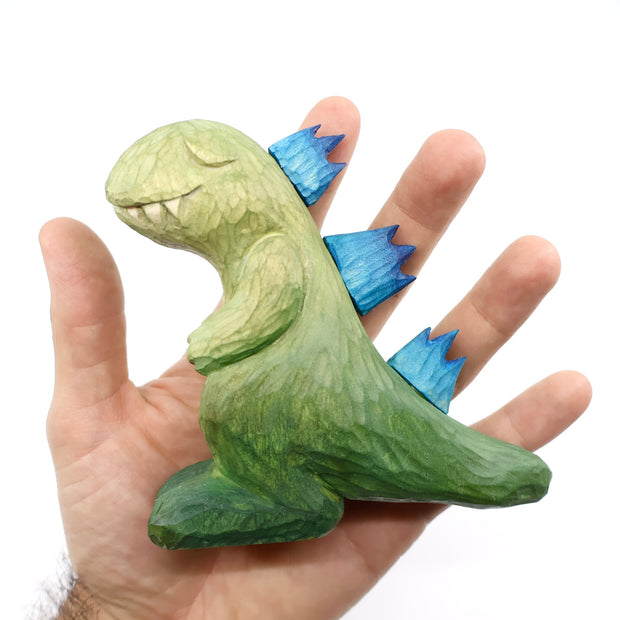 Whittled wooden sculpture of a green Godzilla with blue back spikes, modeled in a non aggressive, cute fashion. Its eyes are peacefully closed and its hands rest on its chest.