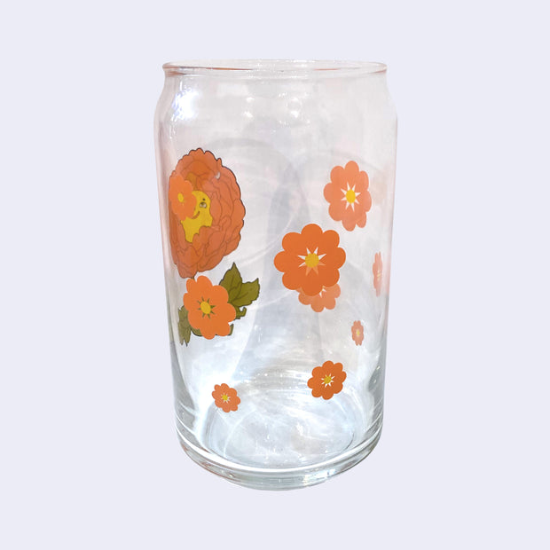 Glass cup with a flat base and slightly inward lip. Features a graphic of a cartoon style orange peony, sitting with a smiling face and a chubby green body made out of stems and leaves. Around the rest of the glass are orange flowers.