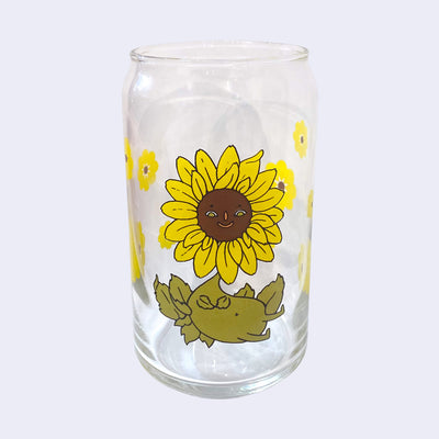 Glass cup with a flat base and slightly inward lip. Features a graphic of a cartoon style sunflower, sitting with a smiling face and a chubby green body made out of stems and leaves. Around the rest of the glass are yellow sunflowers.