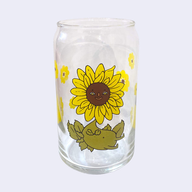 Glass cup with a flat base and slightly inward lip. Features a graphic of a cartoon style sunflower, sitting with a smiling face and a chubby green body made out of stems and leaves. Around the rest of the glass are yellow sunflowers.