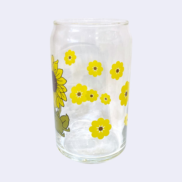Glass cup with a flat base and slightly inward lip. Features a graphic of a cartoon style sunflower, sitting with a smiling face and a chubby green body made out of stems and leaves. Around the rest of the glass are yellow sunflowers. Side view.