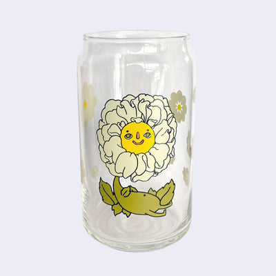 Glass cup with a flat base and slightly inward lip. Features a graphic of a cartoon style white camellia, sitting with a smiling face and a chubby green body made out of stems and leaves. Around the rest of the glass are white flowers.