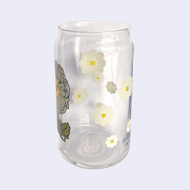 Glass cup with a flat base and slightly inward lip. Features a graphic of a cartoon style white camellia, sitting with a smiling face and a chubby green body made out of stems and leaves. Around the rest of the glass are white flowers. Alt side view.