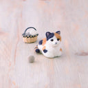 Needle felted calico cat, sitting as a "loaf" with a basket behind it.