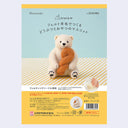 Packaging for a needle felting kit of a polar bear, sitting with a large piece of twisted bread (or a donut twist) in its arms.