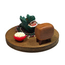 Sculpture of a child in a green kaiju costume laying on a braided carpet and watching a TV, with a bowl of popcorn and a toy robot figurine nearby. A monster movie is on the TV.