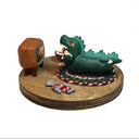 Sculpture of a child in a green kaiju costume laying on a braided carpet and watching a TV, with a bowl of popcorn and a toy robot figurine nearby. A monster movie is on the TV.